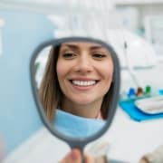 A woman sits in a dental chair, smiling and looking at her reflection in a handheld mirror, with dental tools visible in the background.