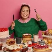 A smiling woman sits at a table filled with assorted desserts, cakes, pastries, and fruit, holding a fork and knife against a pink background.