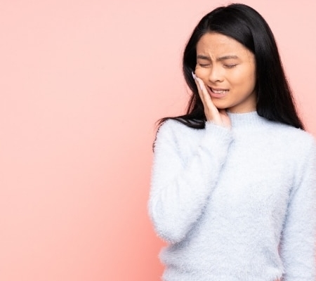 Woman with long black hair touching her cheek and wincing, possibly experiencing tooth or jaw pain, standing against a plain pink background.