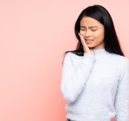 Woman with long black hair touching her cheek and wincing, possibly experiencing tooth or jaw pain, standing against a plain pink background.