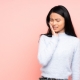 Woman with long black hair touching her cheek and wincing, possibly experiencing tooth or jaw pain, standing against a plain pink background.