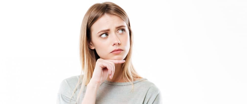 Young woman with long blonde hair wearing a grey shirt, looking up and to the side with a thoughtful expression, hand resting on her chin against a white background.
