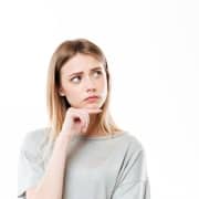 Young woman with long blonde hair wearing a grey shirt, looking up and to the side with a thoughtful expression, hand resting on her chin against a white background.