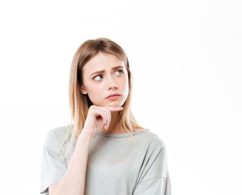 Young woman with long blonde hair wearing a grey shirt, looking up and to the side with a thoughtful expression, hand resting on her chin against a white background.