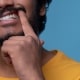 A person in a yellow shirt lifts their upper lip with a finger to show their teeth and gums against a blue background.