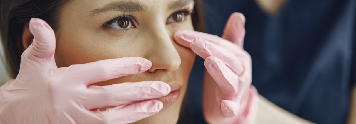 A person wearing pink gloves examines a womans face, gently touching the area under her eyes.