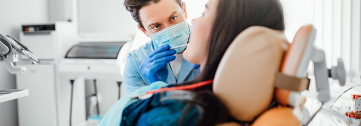 A dentist wearing a mask and gloves examines a patient sitting in a dental chair in a brightly lit clinic.